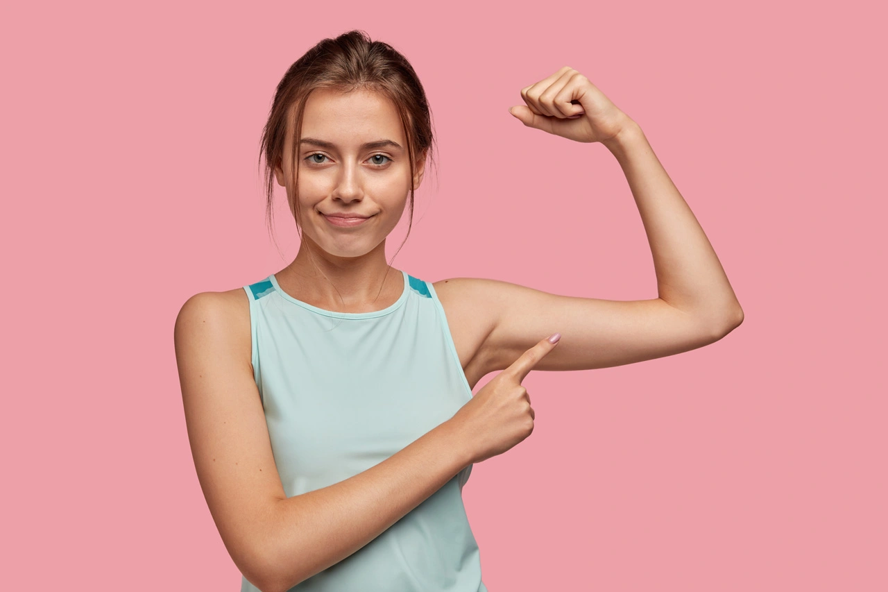 self-assured-pretty-dark-haired-sporty-woman-with-pleasant-appearance-raises-hand-indicates-biceps-confirms-women-can-be-also-strong-wears-light-blue-vest-isolated-pink-wall